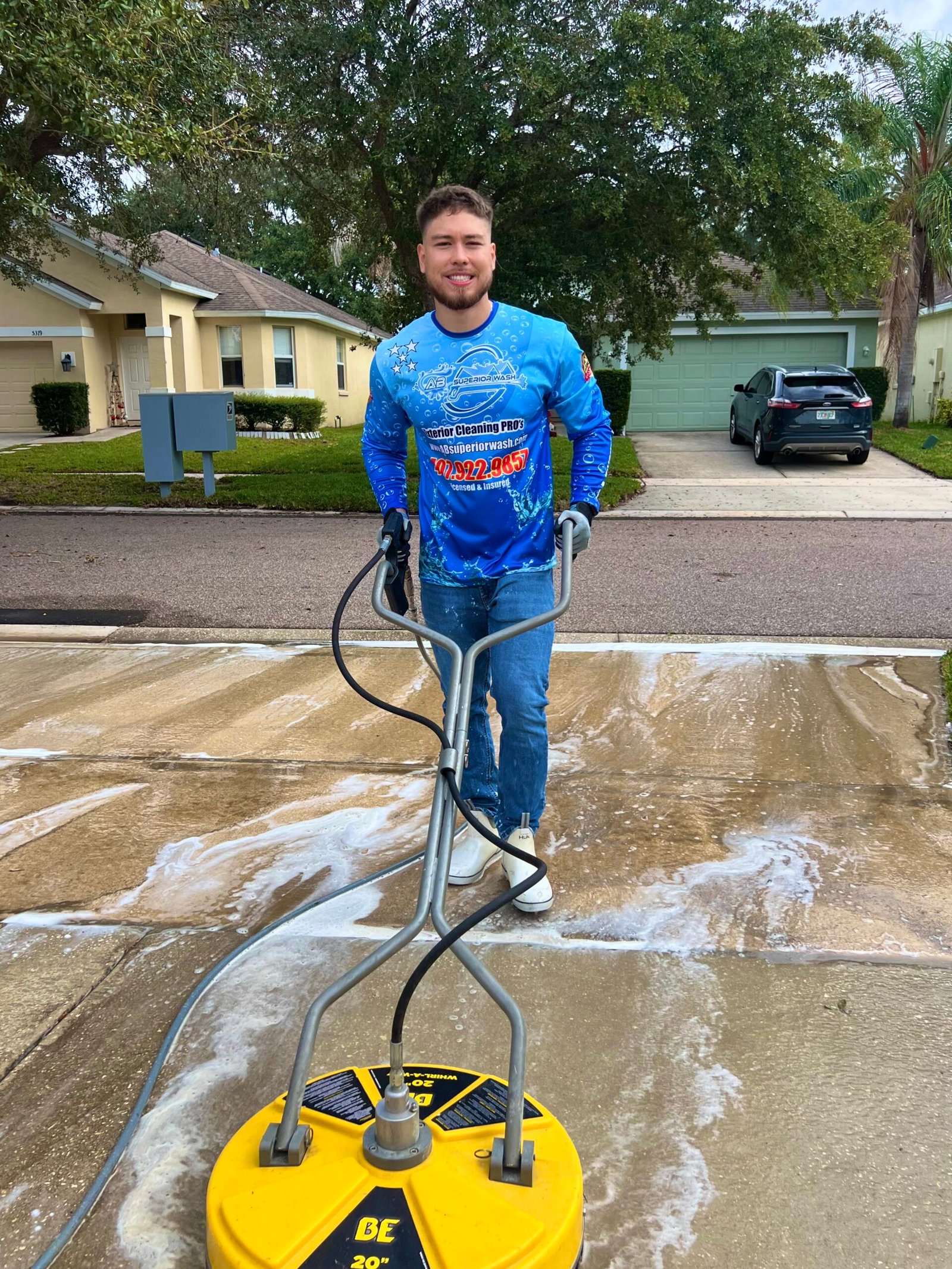 Smiling AB Superior Wash technician in branded jersey with a surface cleaner on a half-cleaned driveway in Orlando, FL